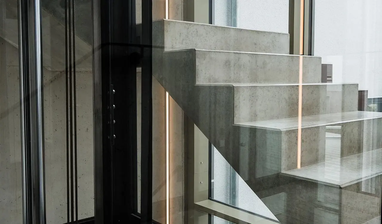 Treppe aus Sichtbeton mit Lichtfuge neben dem gläsernen Aufzug